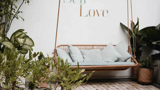 Cozy hanging rattan swing with light blue cushions surrounded by lush green plants and 'Self Love' wall art