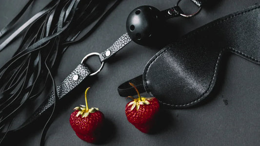 Black leather blindfold, flogger, and gag with fresh strawberries on dark surface